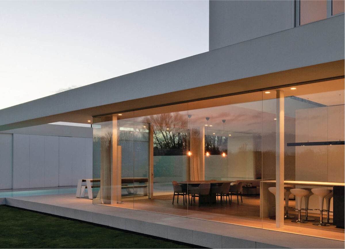 A WinDoor frameless glass wall glowing at dusk on a modern luxury estate — the clean sightlines and expansive openings the brand is specified for.